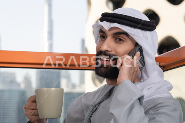 Gestures of happiness and joy, making a phone call, using a modern and advanced technical device, sipping a favorite drink, a close-up image of an Emirati Gulf Arab man wearing a kandura and a white ghutrah holding a mobile phone and a cup of coffee, working remotely, communicating and exchanging conversations with family and friends.