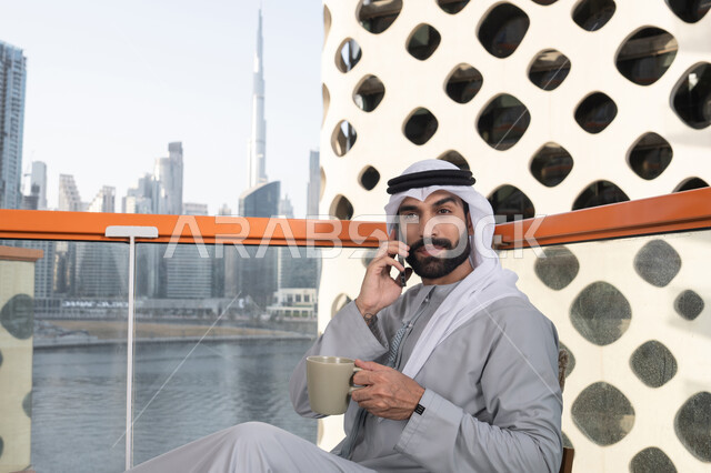 التواصل وتبادل الأحاديث مع الأهل والأصدقاء, اجراء مكالمة هاتفية، استخدام جهاز تقني حديث ومتطور، احتساء المشروب المفضل، رجل عربي خليجي اماراتي يرتدي الكندورة والغترة البيضاء يمسك بيده الهاتف المحمول وكوب من القهوة، العمل عن بعد