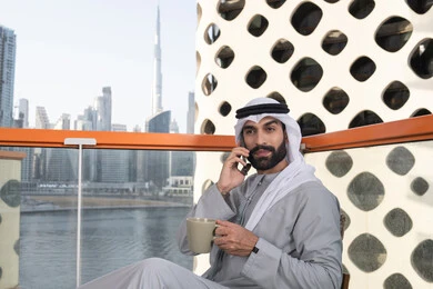 Communication and exchanging conversations with family and friends, making a phone call, using a modern and advanced technical device, sipping a favorite drink, an Emirati Gulf Arab man wearing a kandura and a white ghutrah holding a mobile phone and a cup of coffee, working remotely.