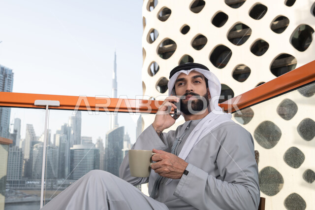 العمل عن بعد, اجراء مكالمة هاتفية، استخدام جهاز تقني حديث ومتطور، احتساء المشروب المفضل، رجل عربي خليجي اماراتي يرتدي الكندورة والغترة البيضاء يمسك بيده الهاتف المحمول وكوب من القهوة، التواصل وتبادل الأحاديث مع الأهل والأصدقاء