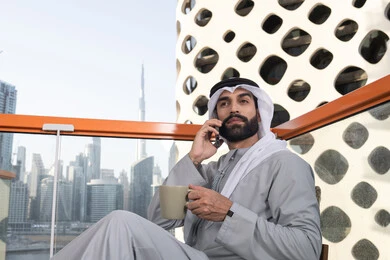 Remote work, making a phone call, using a modern and advanced technical device, sipping a favorite drink, an Emirati Gulf Arab man wearing a kandura and a white ghutrah holding a mobile phone and a cup of coffee, communicating and exchanging conversations with family and friends.