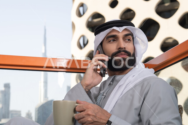 A close-up image of an Emirati Gulf Arab man wearing a kandura and a white ghutrah, holding a mobile phone and a cup of coffee while looking at something. Making a phone call, using a modern and advanced technical device, sipping his favorite drink, working remotely, communicating and exchanging conversations with family and friends.