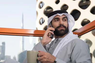 صورة مقربة لرجل عربي خليجي اماراتي يرتدي الكندورة والغترة البيضاء يمسك بيده الهاتف المحمول وكوب من القهوة و ينظر الى شيء ما. اجراء مكالمة هاتفية، استخدام جهاز تقني حديث ومتطور، احتساء المشروب المفضل، العمل عن بعد، التواصل وتبادل الأحاديث مع الأهل والأصدقاء