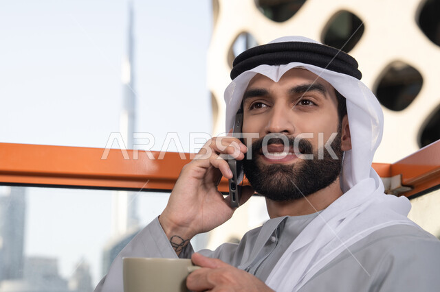 احتساء المشروب المفضل, اجراء مكالمة هاتفية، استخدام جهاز تقني حديث ومتطور، صورة مقربة لرجل عربي خليجي اماراتي يرتدي الكندورة والغترة البيضاء يمسك بيده الهاتف المحمول وكوب من القهوة، العمل عن بعد، التواصل وتبادل الأحاديث مع الأهل والأصدقاء
