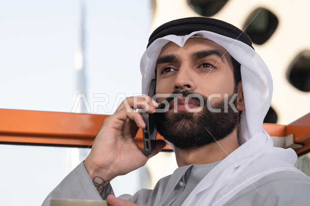 استخدام جهاز تقني حديث ومتطور, اجراء مكالمة هاتفية، احتساء المشروب المفضل، صورة مقربة لرجل عربي خليجي اماراتي يرتدي الكندورة والغترة البيضاء يمسك بيده الهاتف المحمول وكوب من القهوة، العمل عن بعد، التواصل وتبادل الأحاديث مع الأهل والأصدقاء