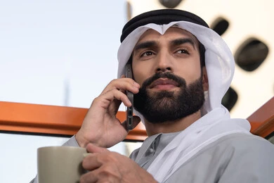 اجراء مكالمة هاتفية، استخدام جهاز تقني حديث ومتطور، احتساء المشروب المفضل، صورة مقربة لرجل عربي خليجي اماراتي يرتدي الكندورة والغترة البيضاء يمسك بيده الهاتف المحمول وكوب من القهوة، العمل عن بعد، التواصل وتبادل الأحاديث مع الأهل والأصدقاء