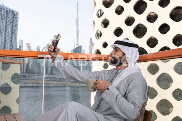 قضاء أوقات ممتعة, توثيق اللحظات السعيدة، التقاط الصور الشخصية باستخدام الجوال، استخدام جهاز تقني حديث ومتطور في تخليد الذكريات، رجل عربي خليجي اماراتي يرتدي الكندورة والغترة البيضاء يمسك الهاتف المحمول ويأخذ صورة سيلفي بإيماءات السرور، احتساء مشروب القهوة