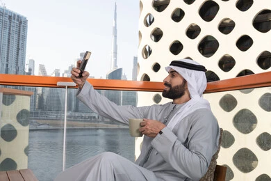 Spending enjoyable times, documenting happy moments, taking selfies using a mobile phone, using a modern and advanced technological device to immortalize memories, an Arab Gulf Emirati man wearing a kandura and a white ghutrah holding a mobile phone and taking a selfie with gestures of joy, sipping a coffee drink.