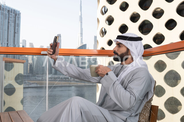 الاستمتاع بشرب المشروبات الساخنه, توثيق اللحظات السعيدة، التقاط الصور الشخصية باستخدام الجوال، استخدام جهاز تقني حديث ومتطور في تخليد الذكريات، رجل عربي خليجي اماراتي يرتدي الكندورة والغترة البيضاء يمسك الهاتف المحمول ويأخذ صورة سيلفي