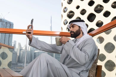 التسلية والترفيه بالعطلات, توثيق اللحظات السعيدة، التقاط الصور الشخصية باستخدام الجوال، استخدام جهاز تقني حديث ومتطور في تخليد الذكريات، رجل عربي خليجي اماراتي يرتدي الكندورة والغترة البيضاء يمسك الهاتف المحمول ويأخذ صورة سيلفي بإيماءات السرور، احتساء مشروب القهوة