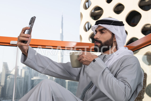 الاستمتاع بقضاء عطله نهاية الاسبوع ,توثيق اللحظات السعيدة، التقاط الصور الشخصية باستخدام الجوال، استخدام جهاز تقني حديث ومتطور في تخليد الذكريات، رجل عربي خليجي اماراتي يرتدي الكندورة والغترة البيضاء يمسك الهاتف المحمول ويأخذ صورة سيلفي بإيماءات السرور، احتساء مشروب القهوة