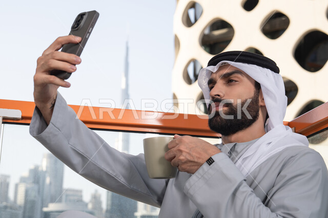 قضاء وقت ممتع فى الخارج,توثيق اللحظات السعيدة، التقاط الصور الشخصية باستخدام الجوال، استخدام جهاز تقني حديث ومتطور في تخليد الذكريات، رجل عربي خليجي اماراتي يرتدي الكندورة والغترة البيضاء يمسك الهاتف المحمول ويأخذ صورة سيلفي بإيماءات السرور، احتساء مشروب القهوة