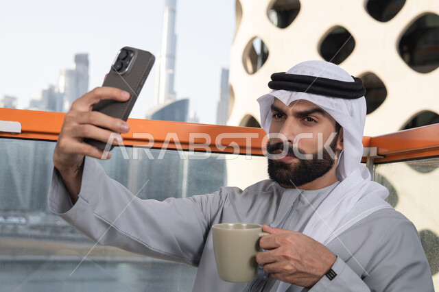 دمج التقنيات والتكنولوجيا الحديثة بالحياة اليومية, توثيق اللحظات السعيدة، التقاط الصور الشخصية باستخدام الجوال، صورة مقربة لرجل عربي خليجي اماراتي يرتدي الكندورة والغترة البيضاء يمسك الهاتف المحمول ويأخذ صورة سيلفي بإيماءات السرور، احتساء مشروب القهوة