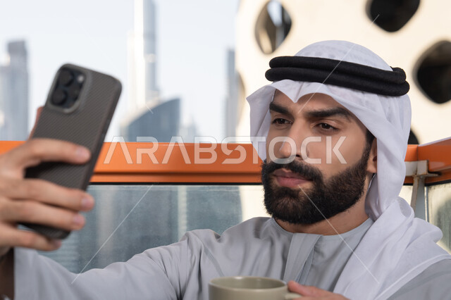 Various mobile programs and applications, documenting happy moments, taking selfies using a mobile phone, using a modern and advanced technological device to immortalize memories, a close-up image of an Emirati Gulf Arab man wearing a kandura and a white ghutrah holding a mobile phone and taking a selfie, sipping a coffee drink.