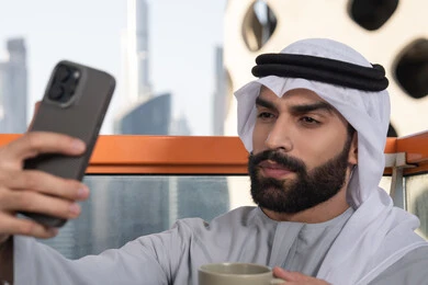 Various mobile programs and applications, documenting happy moments, taking selfies using a mobile phone, using a modern and advanced technological device to immortalize memories, a close-up image of an Emirati Gulf Arab man wearing a kandura and a white ghutrah holding a mobile phone and taking a selfie, sipping a coffee drink.