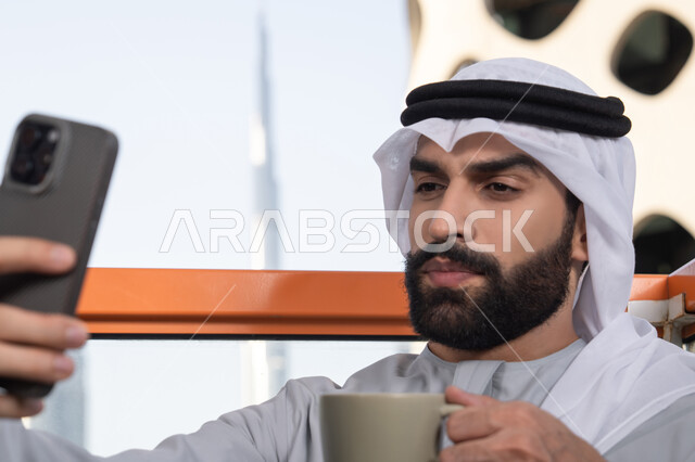 الاستمتاع بتناول المشروبات, توثيق اللحظات السعيدة، التقاط الصور الشخصية باستخدام الجوال، استخدام جهاز تقني حديث ومتطور في تخليد الذكريات،  صورة مقربة لرجل عربي خليجي اماراتي يرتدي الكندورة والغترة البيضاء يمسك الهاتف المحمول ويأخذ صورة سيلفي