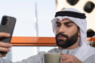 Enjoying drinks, documenting happy moments, taking selfies using a mobile phone, using a modern and advanced technological device to preserve memories, a close-up image of an Emirati Gulf Arab man wearing a kandura and a white ghutrah holding a mobile phone and taking a selfie.