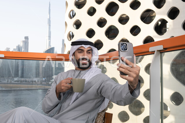 استخدام الأجهزة التقنية الحديثة, توثيق اللحظات السعيدة، التقاط الصور الشخصية باستخدام الجوال، رجل عربي خليجي اماراتي يرتدي الكندورة والغترة البيضاء يمسك الهاتف المحمول ويأخذ صورة سيلفي بإيماءات السرور، احتساء مشروب القهوة