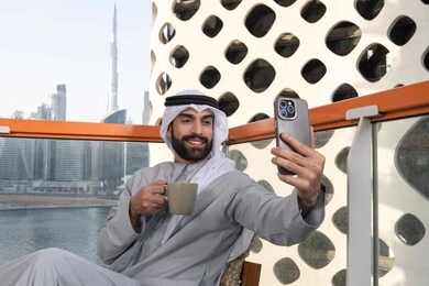 استخدام الأجهزة التقنية الحديثة, توثيق اللحظات السعيدة، التقاط الصور الشخصية باستخدام الجوال، رجل عربي خليجي اماراتي يرتدي الكندورة والغترة البيضاء يمسك الهاتف المحمول ويأخذ صورة سيلفي بإيماءات السرور، احتساء مشروب القهوة