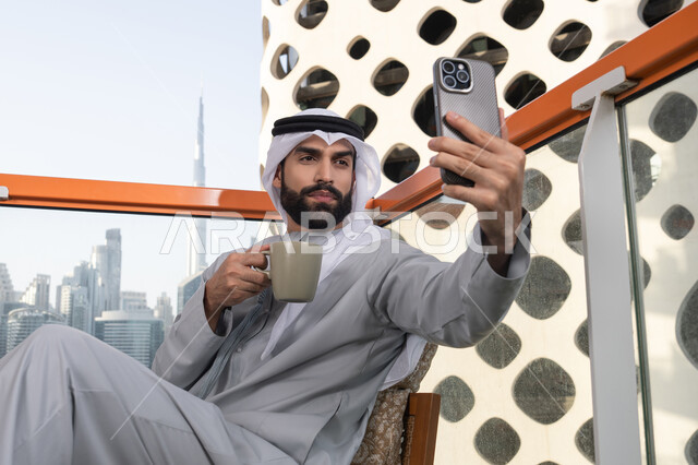 الاحتفاظ باللحظات السعيدة، التقاط الصور الشخصية باستخدام الجوال، استخدام جهاز تقني حديث ومتطور في تخليد الذكريات، رجل عربي خليجي اماراتي يرتدي الكندورة والغترة البيضاء يمسك الهاتف المحمول ويأخذ صورة سيلفي بإيماءات السرور، احتساء مشروب القهوة