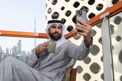 الاحتفاظ باللحظات السعيدة، التقاط الصور الشخصية باستخدام الجوال، استخدام جهاز تقني حديث ومتطور في تخليد الذكريات، رجل عربي خليجي اماراتي يرتدي الكندورة والغترة البيضاء يمسك الهاتف المحمول ويأخذ صورة سيلفي بإيماءات السرور، احتساء مشروب القهوة