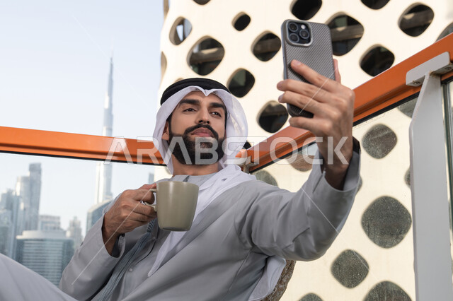 احتساء مشروب القهوة, توثيق اللحظات السعيدة، التقاط الصور الشخصية باستخدام الجوال، استخدام جهاز تقني حديث ومتطور في تخليد الذكريات، رجل عربي خليجي اماراتي يرتدي الكندورة والغترة البيضاء يمسك الهاتف المحمول ويأخذ صورة سيلفي بإيماءات السرور
