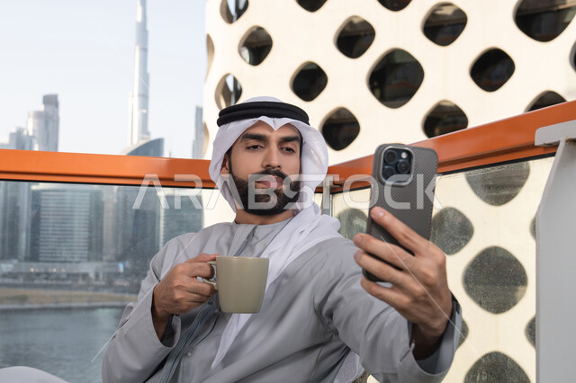 رجل عربي خليجي اماراتي يرتدي الكندورة والغترة البيضاء يمسك الهاتف المحمول ويأخذ صورة سيلفي بإيماءات السرور، توثيق اللحظات السعيدة، التقاط الصور الشخصية باستخدام الجوال، استخدام جهاز تقني حديث ومتطور في تخليد الذكريات، احتساء مشروب القهوة