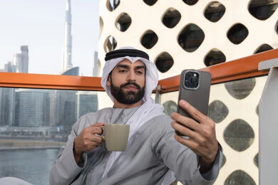 رجل عربي خليجي اماراتي يرتدي الكندورة والغترة البيضاء يمسك الهاتف المحمول ويأخذ صورة سيلفي بإيماءات السرور، توثيق اللحظات السعيدة، التقاط الصور الشخصية باستخدام الجوال، استخدام جهاز تقني حديث ومتطور في تخليد الذكريات، احتساء مشروب القهوة