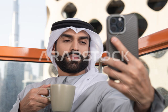 استخدام جهاز تقني حديث ومتطور في تخليد الذكريات, توثيق اللحظات السعيدة، التقاط الصور الشخصية باستخدام الجوال،صورة مقربة لرجل عربي خليجي اماراتي يرتدي الكندورة والغترة البيضاء يمسك الهاتف المحمول ويأخذ صورة سيلفي بإيماءات السرور، احتساء مشروب القهوة