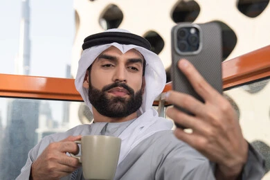 The use of a modern and advanced technical device to immortalize memories, document happy moments, and take personal photos using a mobile phone. A close-up image of an Emirati Gulf Arab man wearing a kandura and a white ghutrah holding a mobile phone and taking a selfie with gestures of joy, sipping a coffee drink.