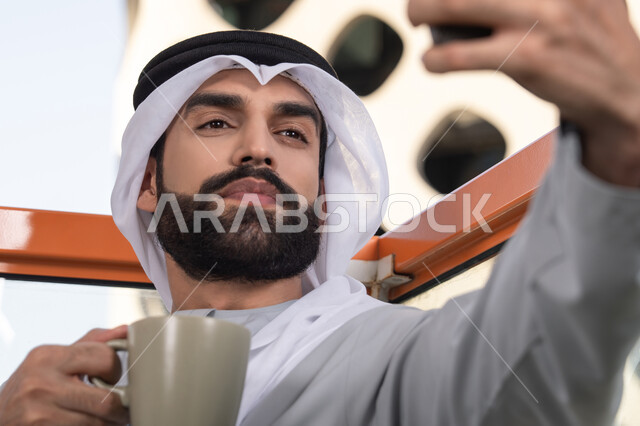Taking selfies using a mobile phone, documenting happy moments, using a modern and advanced technological device to immortalize memories, a close-up image of an Emirati Gulf Arab man wearing a kandura and a white ghutrah holding a mobile phone and taking a selfie with gestures of joy, sipping a coffee drink.