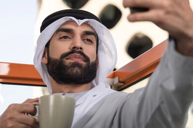 Taking selfies using a mobile phone, documenting happy moments, using a modern and advanced technological device to immortalize memories, a close-up image of an Emirati Gulf Arab man wearing a kandura and a white ghutrah holding a mobile phone and taking a selfie with gestures of joy, sipping a coffee drink.