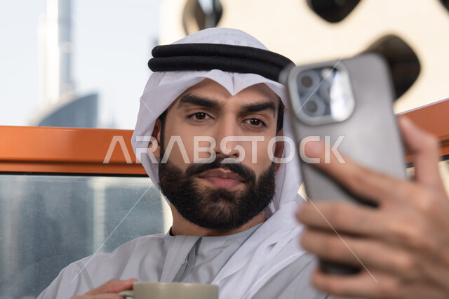 Documenting happy moments, taking selfies using a mobile phone, using a modern and advanced technological device to immortalize memories, a close-up image of an Emirati Gulf Arab man wearing a kandura and a white ghutrah holding a mobile phone and taking a selfie with gestures of joy, sipping a coffee drink.