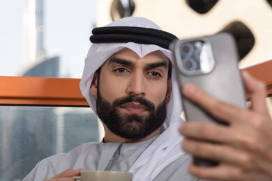 Documenting happy moments, taking selfies using a mobile phone, using a modern and advanced technological device to immortalize memories, a close-up image of an Emirati Gulf Arab man wearing a kandura and a white ghutrah holding a mobile phone and taking a selfie with gestures of joy, sipping a coffee drink.