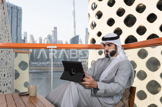 تصفح مواقع التواصل الاجتماعي، إدارة شؤون الأعمال وتنظيمها عبر جهاز التابلت، دمج العمل مع التقنية والتكنولوجيا ، شاب عربي خليجي إماراتي يرتدي الكندورة والغترة البيضاء يجلس في المقهى، قضاء وقت ممتع فى الخارج