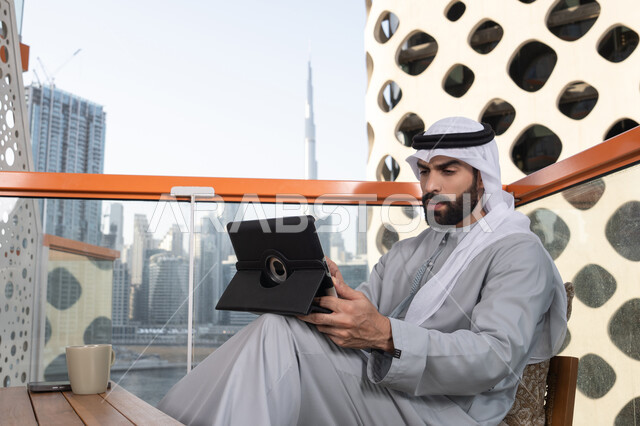 Following news on social media, using a tablet to accomplish projects and tasks, the concept of technology and using modern techniques, a young Arab Gulf Emirati man wearing a kandura and a white ghutrah sitting in a café, spending enjoyable time outdoors.