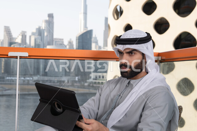 إجراء مكالمات الفيديو مع الأهل والأصدقاء عبر الإنترنت، استخدم جهاز التابلت لانجاز المشاريع والمهام،  مفهوم التكنولوجيا وإستخدام تقنيات حديثة ، شاب عربي خليجي إماراتي يرتدي الكندورة والغترة البيضاء يجلس في المقهى، قضاء وقت ممتع فى الخارج