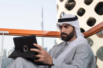 Exchanging conversations, communication, and news with family and friends, using a tablet to accomplish projects and tasks, the concept of technology and the use of modern techniques, a young Arab Gulf Emirati man wearing a kandura and a white ghutrah sitting in a café, spending enjoyable time outdoors.