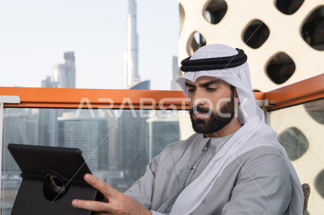 استخدم جهاز التابلت لانجاز المشاريع والمهام،  مفهوم التكنولوجيا وإستخدام تقنيات حديثة ،صورة مقربه لشاب عربي خليجي إماراتي يرتدي الكندورة والغترة البيضاء يجلس في المقهى، قضاء وقت ممتع فى الخارج ، اجراء مكالمه فيديو