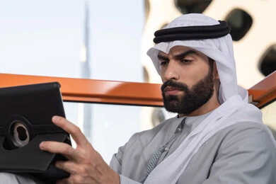 The concept of technology and the use of modern techniques, a close-up image of a young Arab Gulf Emirati man wearing a kandura and a white ghutrah sitting in a café, enjoying time outdoors, making a video call, using a tablet to accomplish projects and tasks.