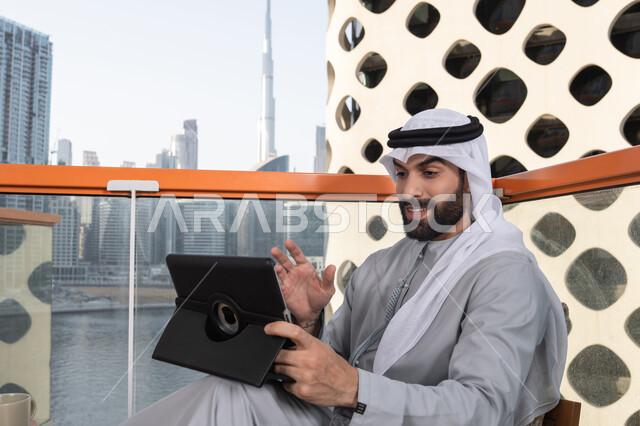 شاب عربي خليجي إماراتي يرتدي الكندورة والغترة البيضاء يجلس في المقهى، قضاء وقت ممتع فى الخارج ، اجراء مكالمه فيديو، استخدم جهاز التابلت لانجاز المشاريع والمهام ، مفهوم التكنولوجيا وإستخدام تقنيات حديثة