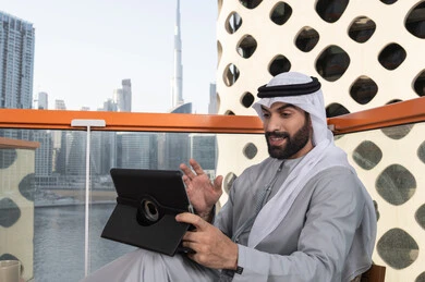 A young Emirati Arab man wearing a kandura and a white ghutrah is sitting in a café, enjoying his time outdoors, making a video call, using a tablet to accomplish projects and tasks, understanding technology and using modern techniques.