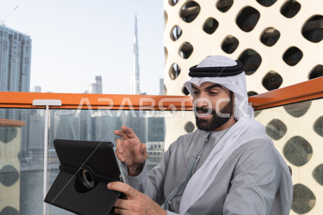 قضاء وقت ممتع فى الخارج ، اجراء مكالمه فيديو، استخدم جهاز التابلت لانجاز المشاريع والمهام ، مفهوم التكنولوجيا وإستخدام تقنيات حديثة، شاب عربي خليجي إماراتي يرتدي الكندورة والغترة البيضاء يجلس في المقهى