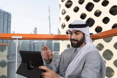 قضاء وقت ممتع فى الخارج ، اجراء مكالمه فيديو، استخدم جهاز التابلت لانجاز المشاريع والمهام ، مفهوم التكنولوجيا وإستخدام تقنيات حديثة، شاب عربي خليجي إماراتي يرتدي الكندورة والغترة البيضاء يجلس في المقهى