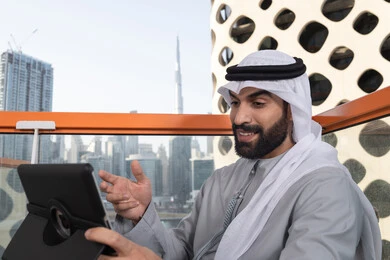 Remote work, using a tablet to complete projects in a café, making a video call via modern technological devices, an Arab Gulf Emirati man wearing a kandura and a white ghutrah sitting in a luxurious coffee shop in the UAE.