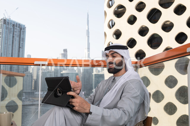 استخدام التكنولوجيا والتقنيات الحديثه، رجل عربي خليجي اماراتي مبتسم يرتدي الكندورة والغترة البيضاء يجلس في كوفي شوب راقي بالامارات ، العمل عن بعد، استخدام جهاز التابلت لانجاز المشاريع في المقهي
