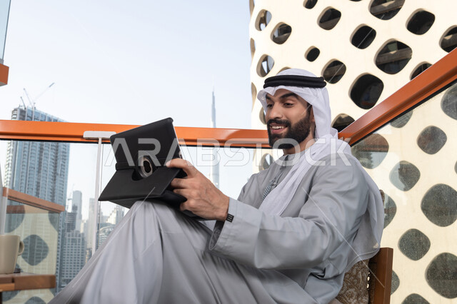 اجراء مكالمة فيديو عبر الاجهزة التقنية الحديثة، كوفي شوب راقي بالامارات، دمج التقنية والتكنولوجيا في الروتين اليومي، تعابير السعادة و السرور، رجل عربي خليجي اماراتي يرتدي الكندورة و الغتره  يستخدم التابلت في مقهى مرتفع مطل على المدينة