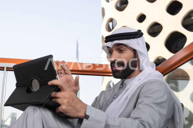 اجراء مكالمه فيديو، كوفي شوب راقي بالامارات، دمج التقنية والتكنولوجيا في الروتين اليومي، تعابير السعادة و السرور، رجل عربي خليجي اماراتي يرتدي الكندورة و الغتره  يستخدم التابلت في مقهى مرتفع مطل على المدينة
