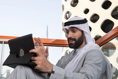 Making a video call, a high-end coffee shop in the UAE, integrating technology and tech into daily routines, expressions of happiness and joy, an Arab Gulf Emirati man wearing a kandura and ghutrah using a tablet in an elevated café overlooking the city.