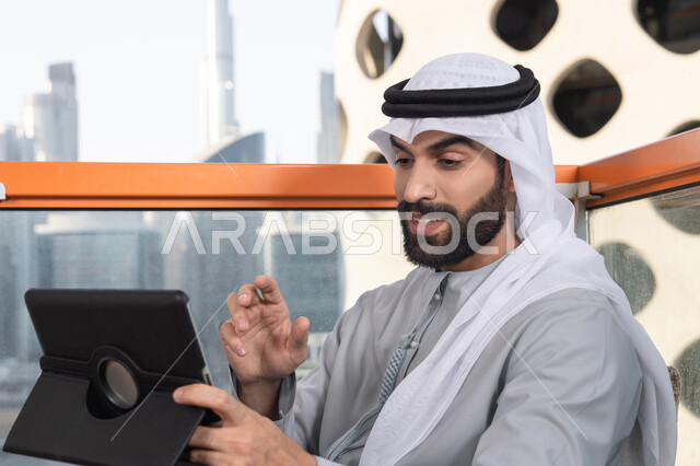 كوفي شوب راقي بالامارات، دمج التقنية والتكنولوجيا في الروتين اليومي، تعابير السعادة و السرور،  صورة مقربه لرجل عربي خليجي اماراتي يرتدي الكندورة و الغتره  يستخدم التابلت في مقهى مرتفع مطل على المدينة